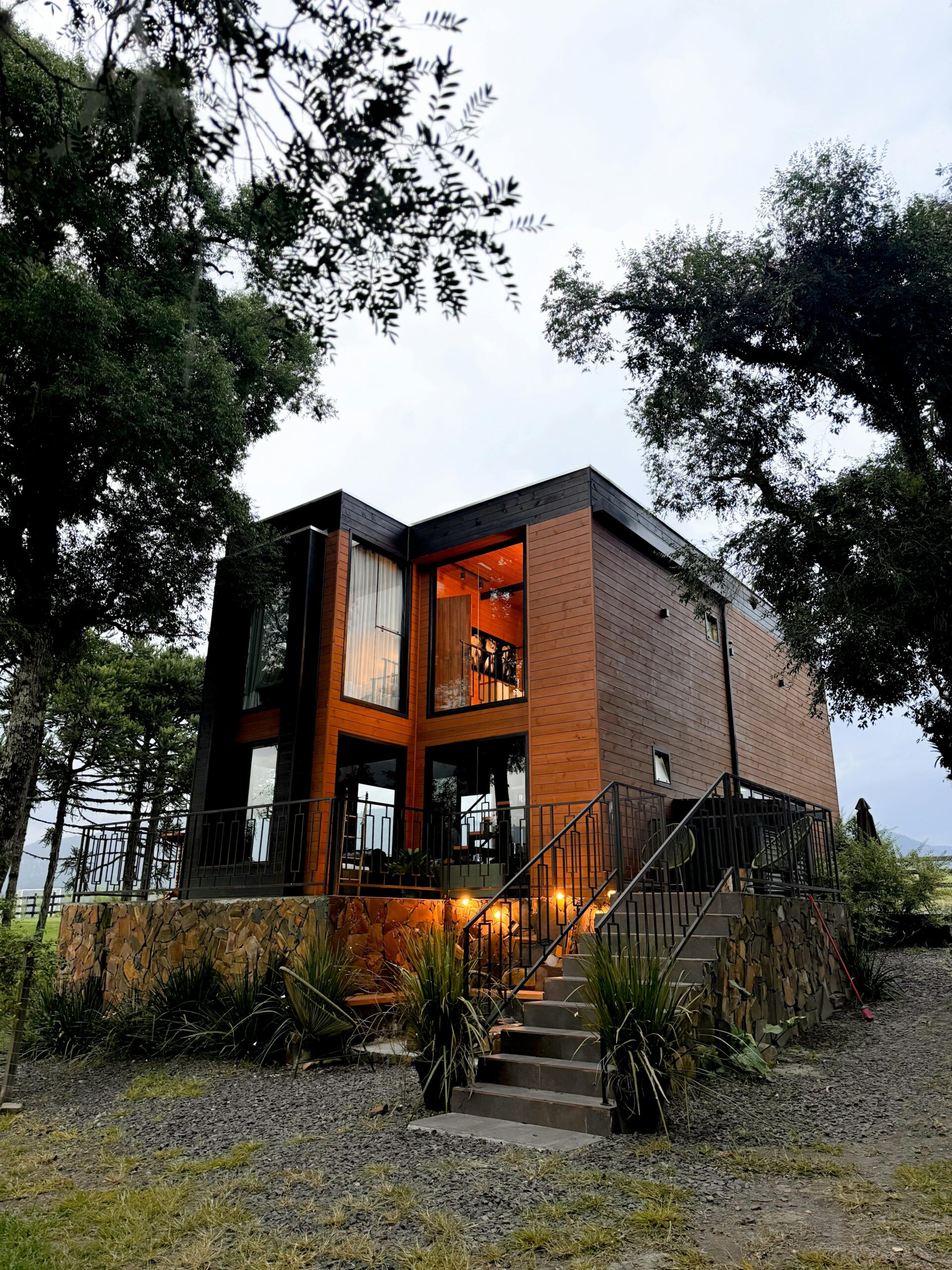 Fotografía de una casa modular de lujo de dos plantas terminada, con fachada de madera tecnológica y grandes ventanales, ubicada sobre una base de piedra natural y rodeada de vegetación, mostrando el diseño moderno y eficiente de Luxury Family Homes.
